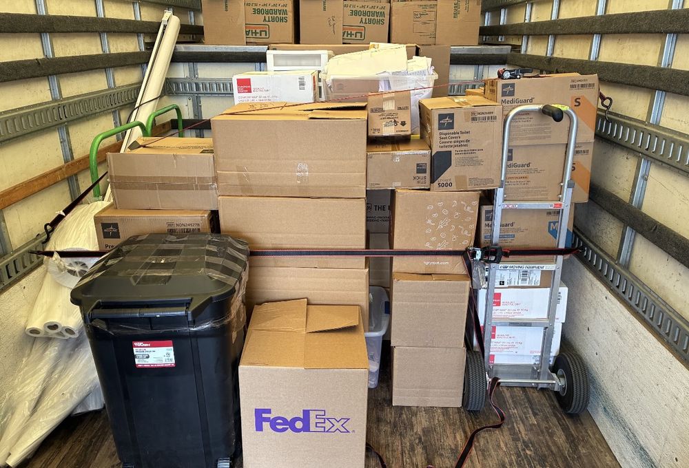Cartons and supply boxes stacked in tight rows inside a truck and secured with a load strap, with a hand truck alongside