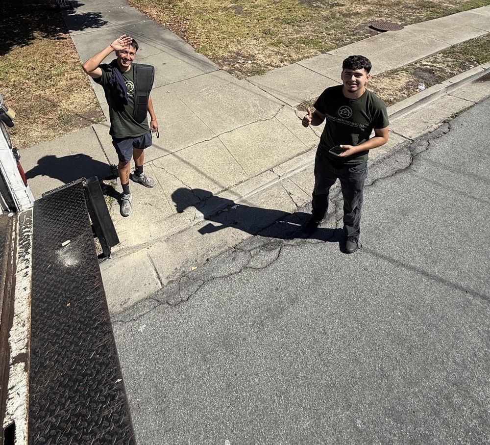 Two Working Men Moving crew members in green company shirts smiling and waving from the sidewalk beside the truck
