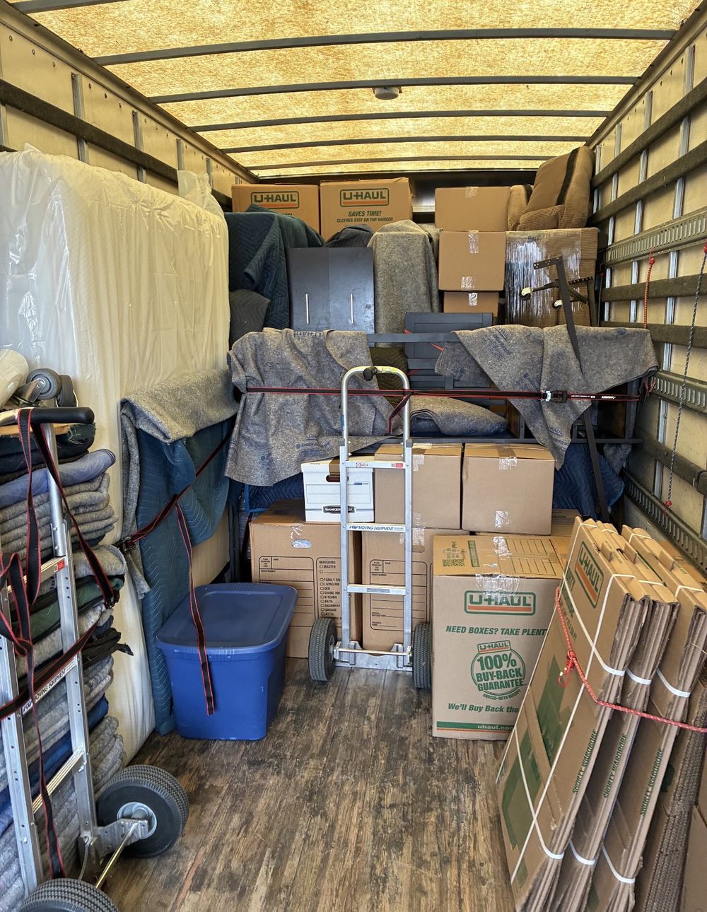 Inside a moving truck: furniture wrapped in moving blankets, labeled boxes stacked neatly and strapped in place, with hand trucks secured along the wall